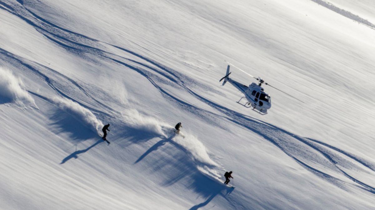 Three snowboarders carve tracks in fresh snow, with a helicopter hovering in the background above a white landscape.
