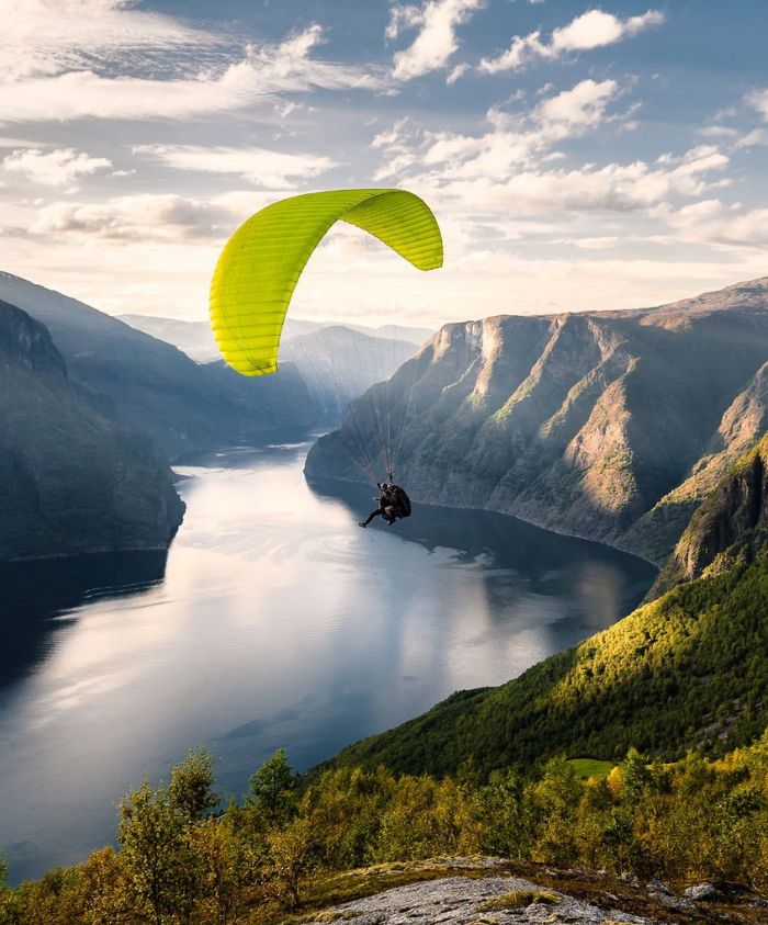 A paraglider with a bright green wing glides over a serene fjord surrounded by steep mountains, reflecting the sky's colors in the calm water below.