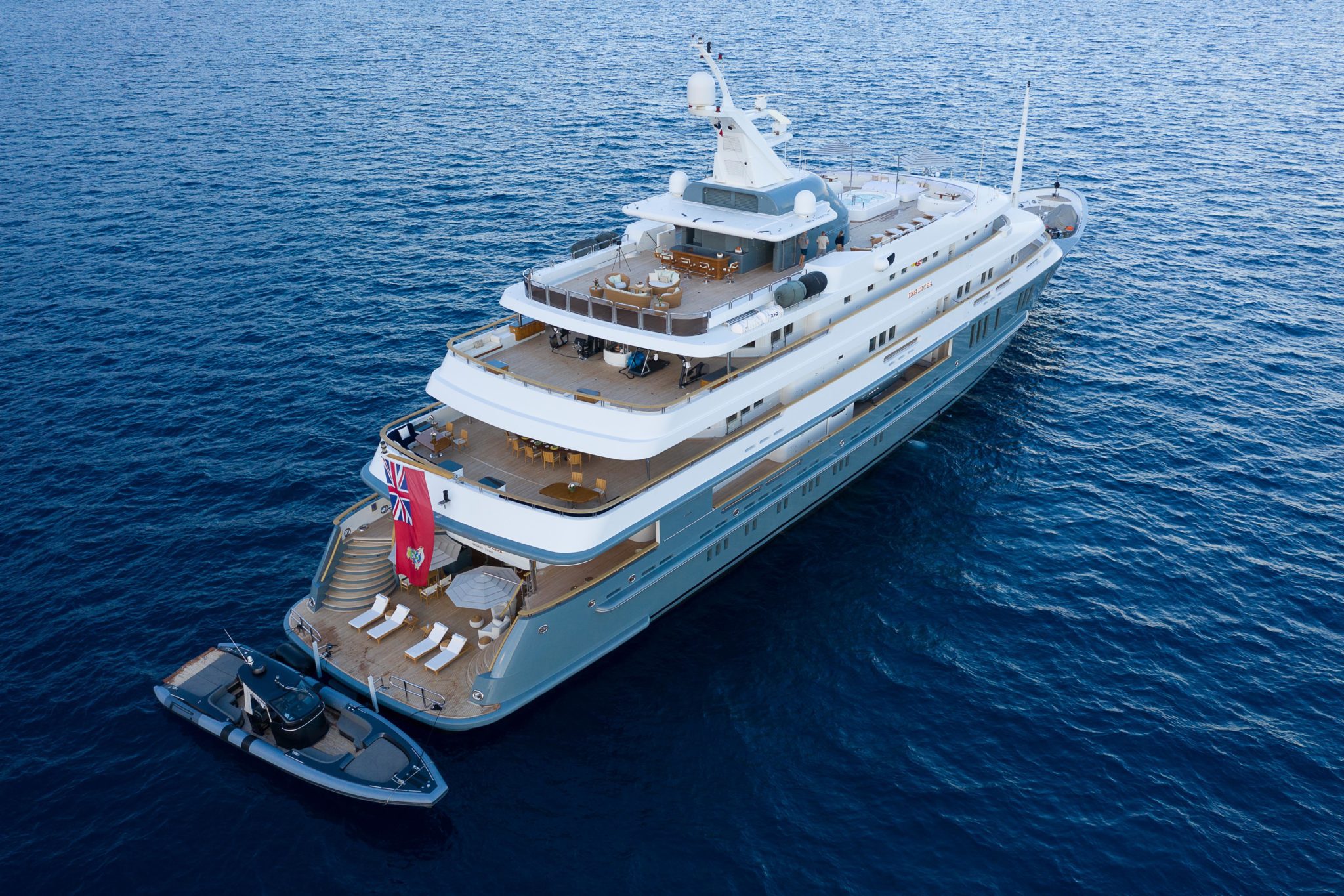 A large luxury yacht anchored in deep blue water, featuring multiple decks with sun loungers and an upper deck dining area. A smaller boat is tied alongside.
