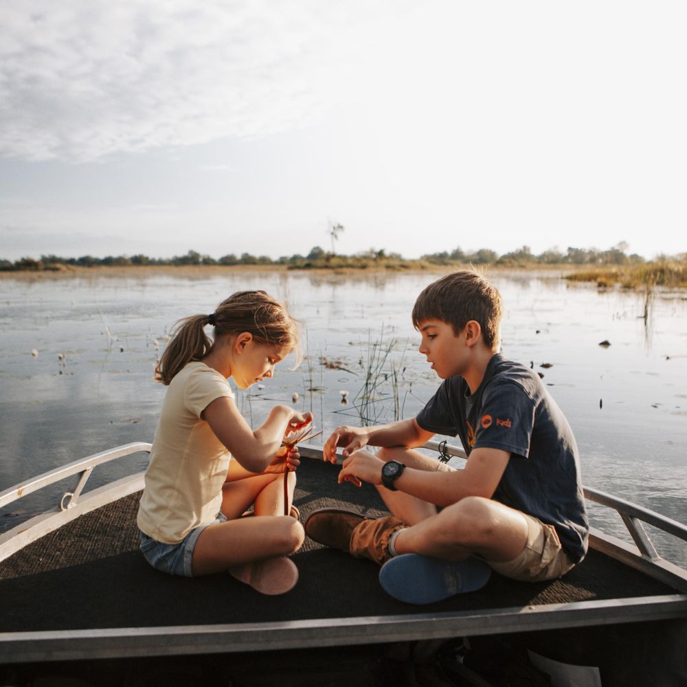 Two children sitting on a boat deck, focused on a task together. The boy is holding something in his hands while the girl is examining it. The background features a serene water landscape with lush greenery and a cloudy sky.