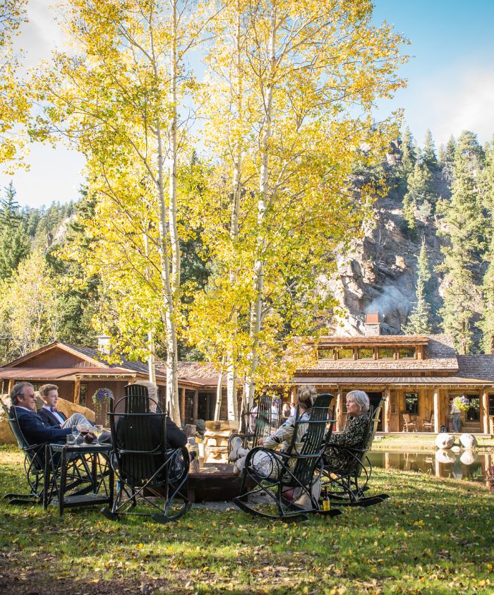 A group of people relaxes in rocking chairs around a fire pit, surrounded by autumn trees. A rustic lodge is visible in the background amid a mountainous landscape.
