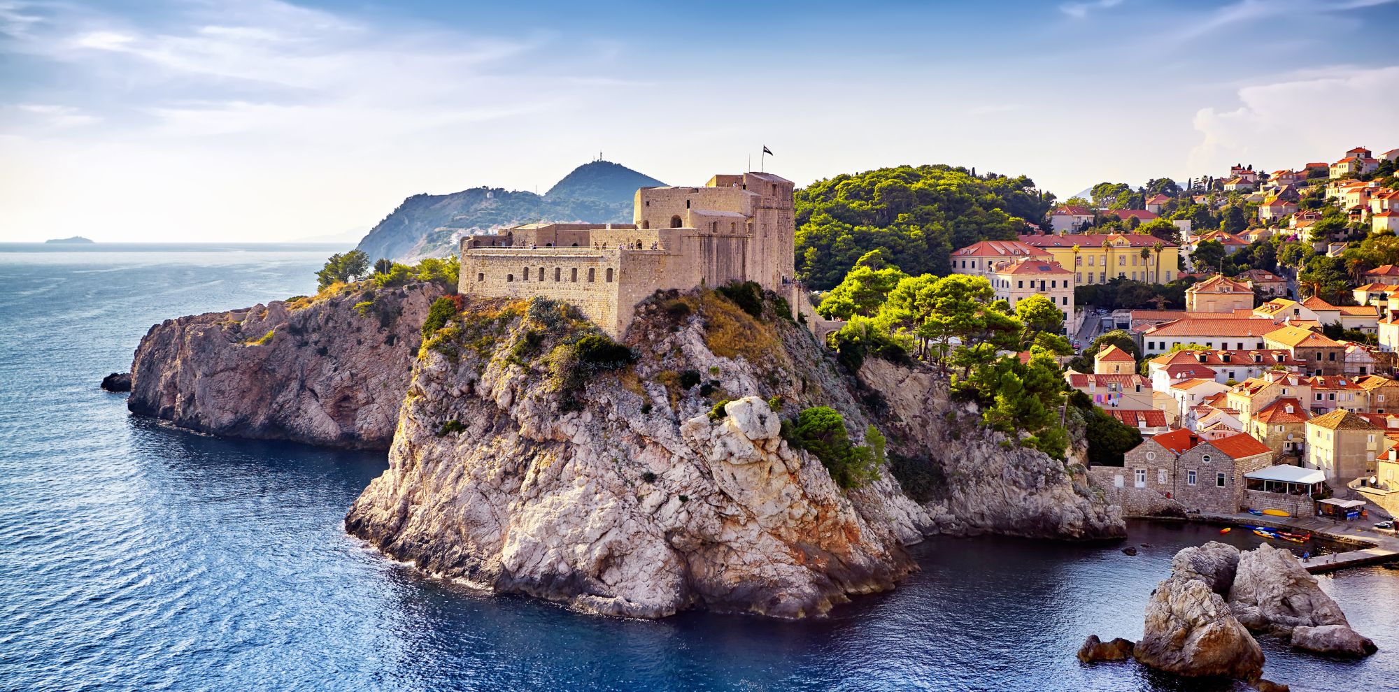 A scenic view of a historic castle perched on a rocky cliff overlooking the turquoise sea, with colorful buildings and lush greenery in the background. The sky is partly cloudy, adding to the picturesque landscape.