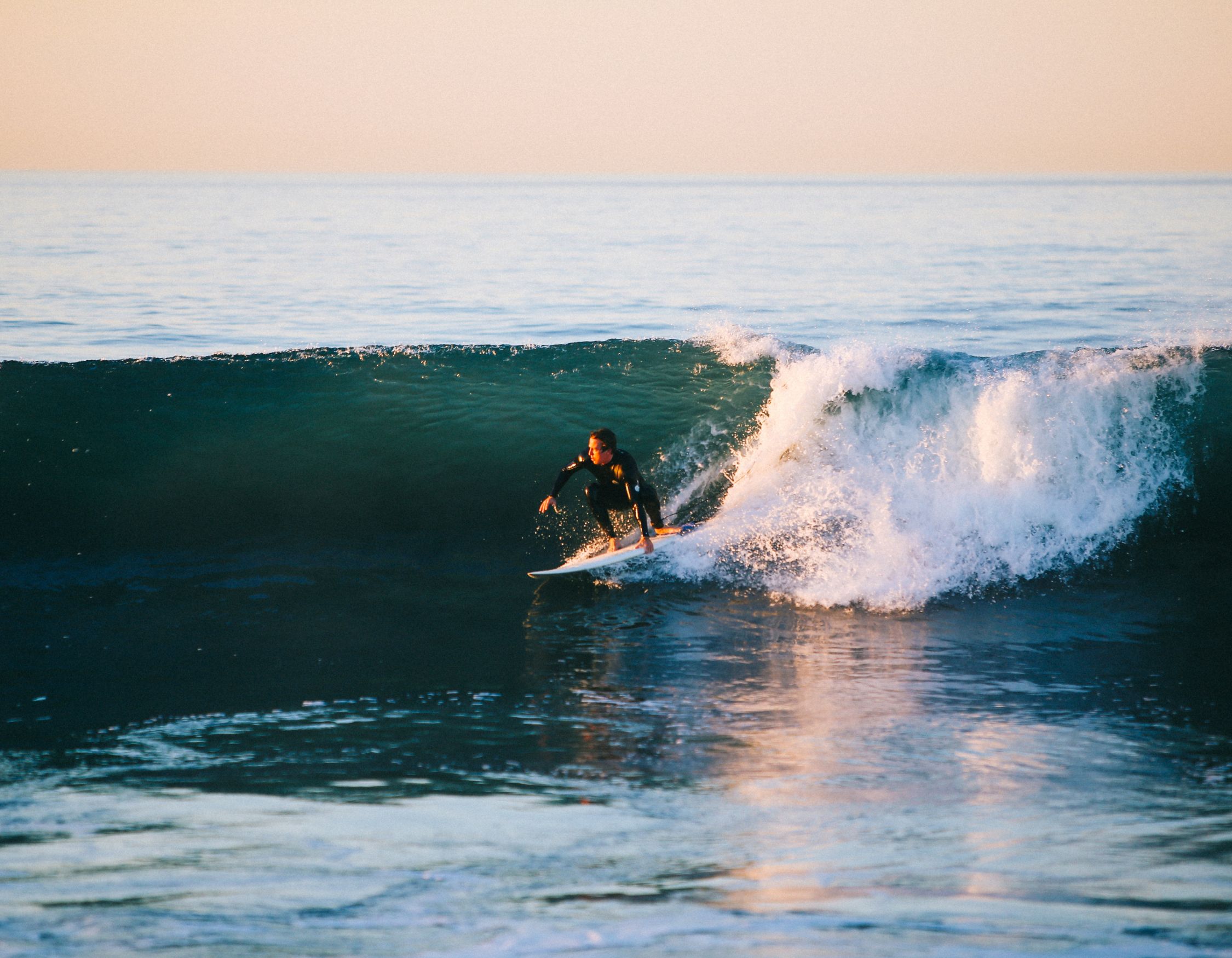 A surfer rides a large wave at sunrise, gliding smoothly on the water while spray and foam splash around. The ocean is calm with a pastel sky in the background.