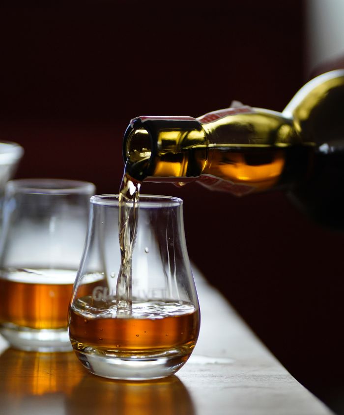 A bottle pouring amber liquid into a curved glass, with two additional glasses in the background. The setting is warm and inviting, suggesting a cozy atmosphere.