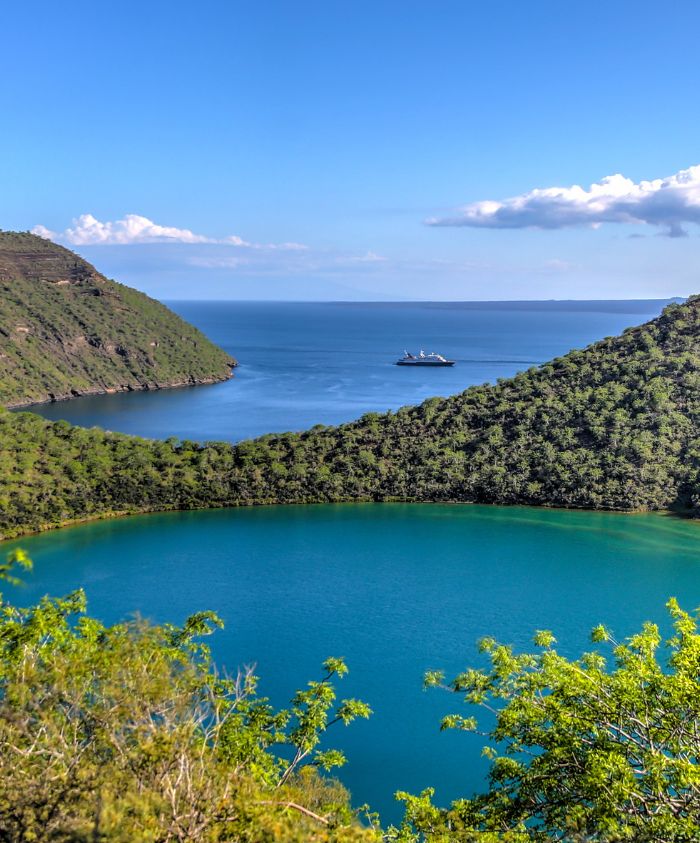 Aerial view of untouched Galápagos Islands surrounded by turquoise waters and a luxury superyacht in the background, Pelorus Yachting 2026 luxury expedition yacht.