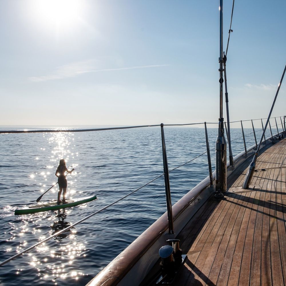 Side view of sailing yacht with paddleboarder gliding beside in turquoise Mediterranean waters – Pelorus Yachting 2026 luxury Odyssey trip.