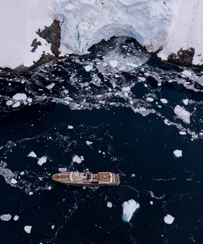 Aerial view of luxury superyacht cruising beneath towering Greenland cliffs – Pelorus Yachting 2026 Arctic exploration voyage.